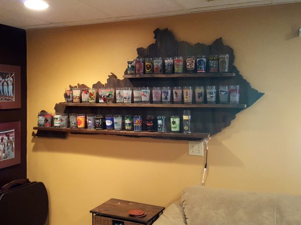 Backlit Handcrafted Barnwood Kentucky Derby Glass Shelf with glasses hanging slightly slanted to the left on a wall.