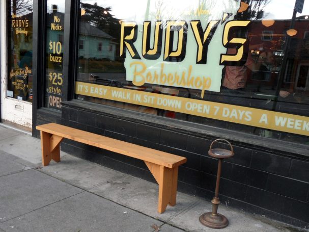 Commercial quality eco-friendly Outdoor Farmhouse Bench slanted to the left on the sidewalk outside a barbershop.