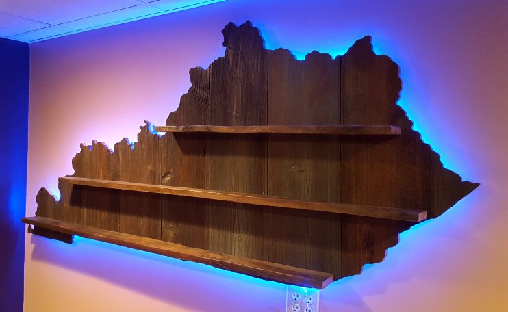 Backlit Handcrafted Barnwood Kentucky Derby Glass Shelf without glasses hanging slanted to the left on a yellow wall. 