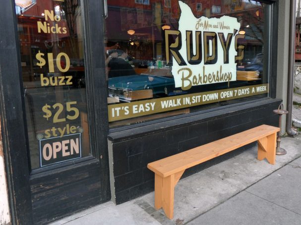 Commercial quality eco-friendly Outdoor Farmhouse Bench slanted to the right on the sidewalk outside a barbershop.