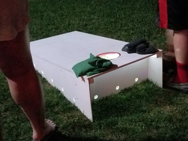 Back of a sturdy high quality solid box design Cornhole Board slanted left in a yard over a light to play at night.