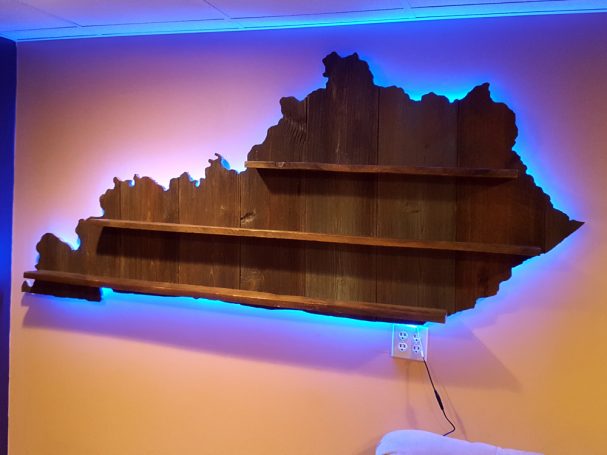 Backlit Handcrafted Barnwood Kentucky Derby Glass Shelf without glasses hanging slightly slanted to the left on a wall. 