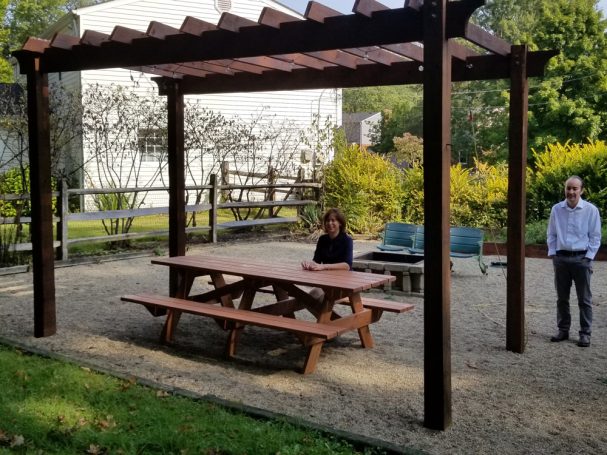 Commercial quality eco-friendly Outdoor Attached Bench Picnic Table slanted to the left on a patio under a pergola.